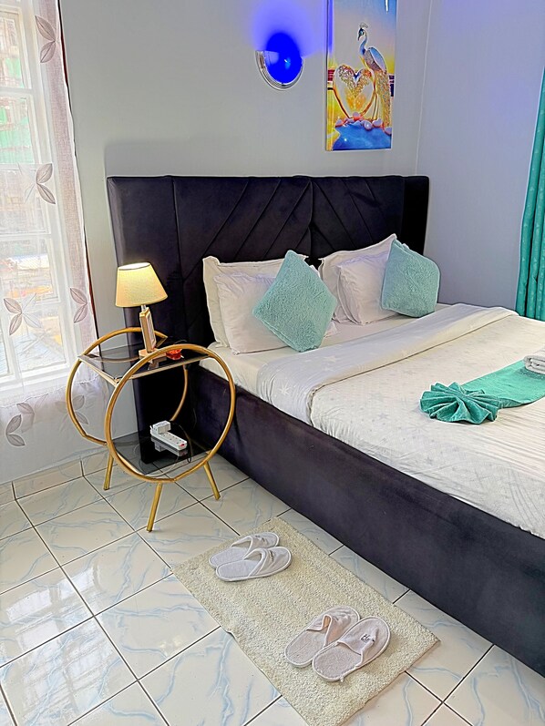 Un dormitorio con cama, mesita de noche con lámpara, decoración en la pared y ventana con cortinas.