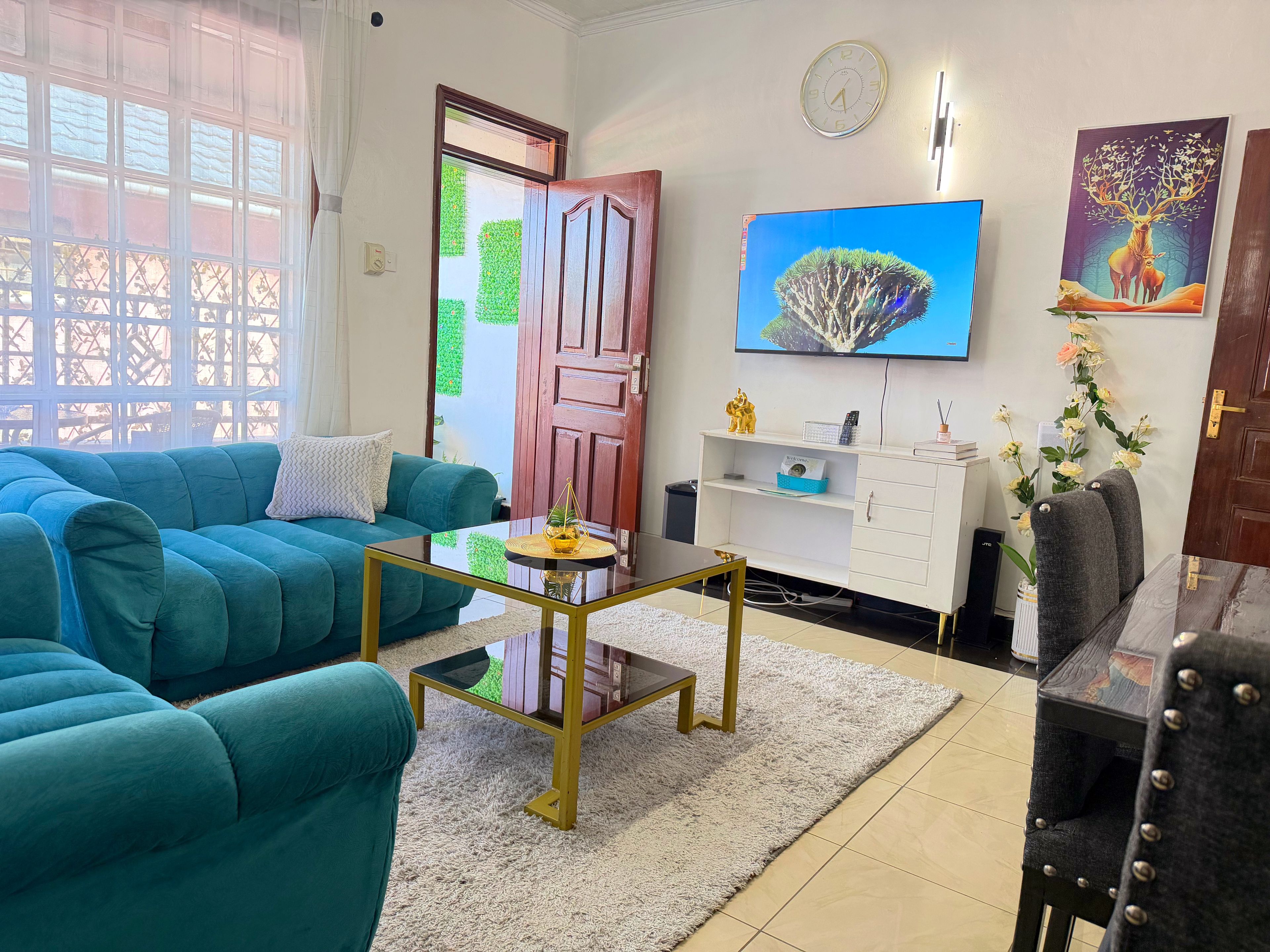 Ein Wohnzimmer mit petrolfarbenen Sofas, einem Glastisch, einem weißen Schrank und einem an der Wand befestigten Fernseher.