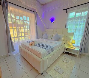 A bedroom with a large bed, bedside tables, a lamp, and curtains. - Homely apartment 2 (Kisumu)