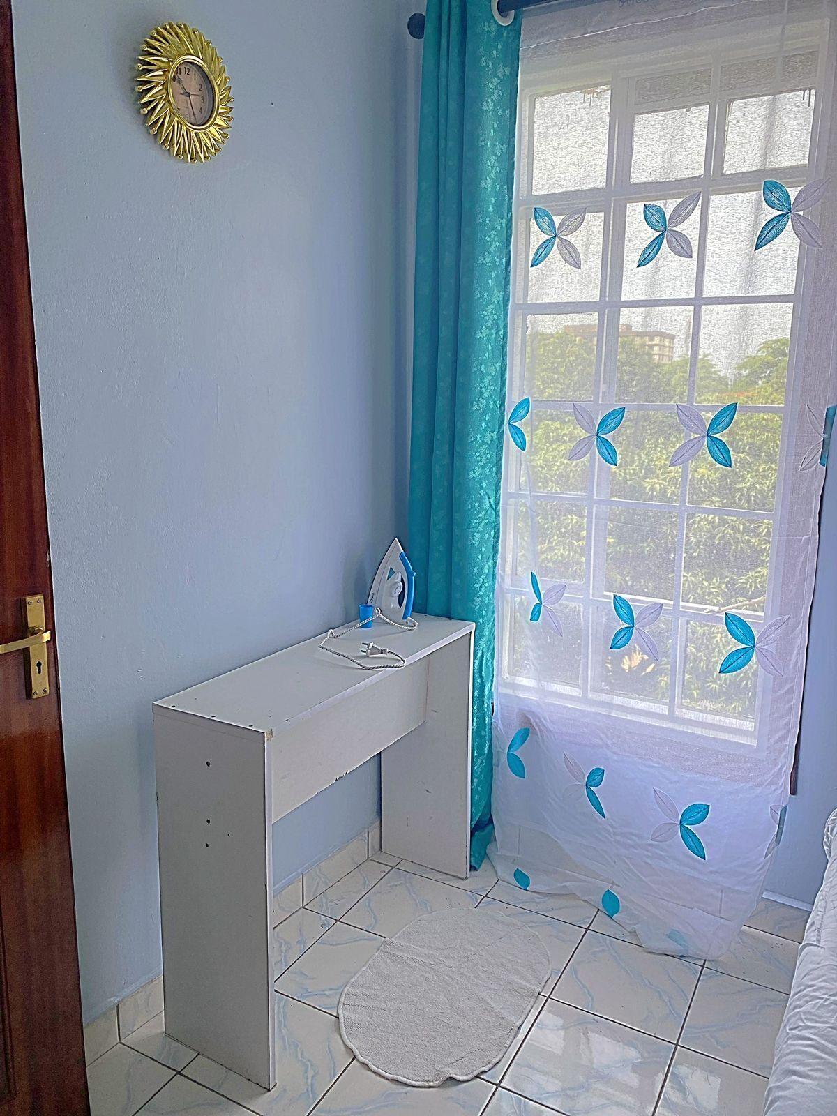 A small, tidy room with a desk, a window with blue curtains, and a decorative sun on the wall.