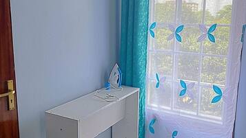 A small, tidy room with a desk, a window with blue curtains, and a decorative sun on the wall.