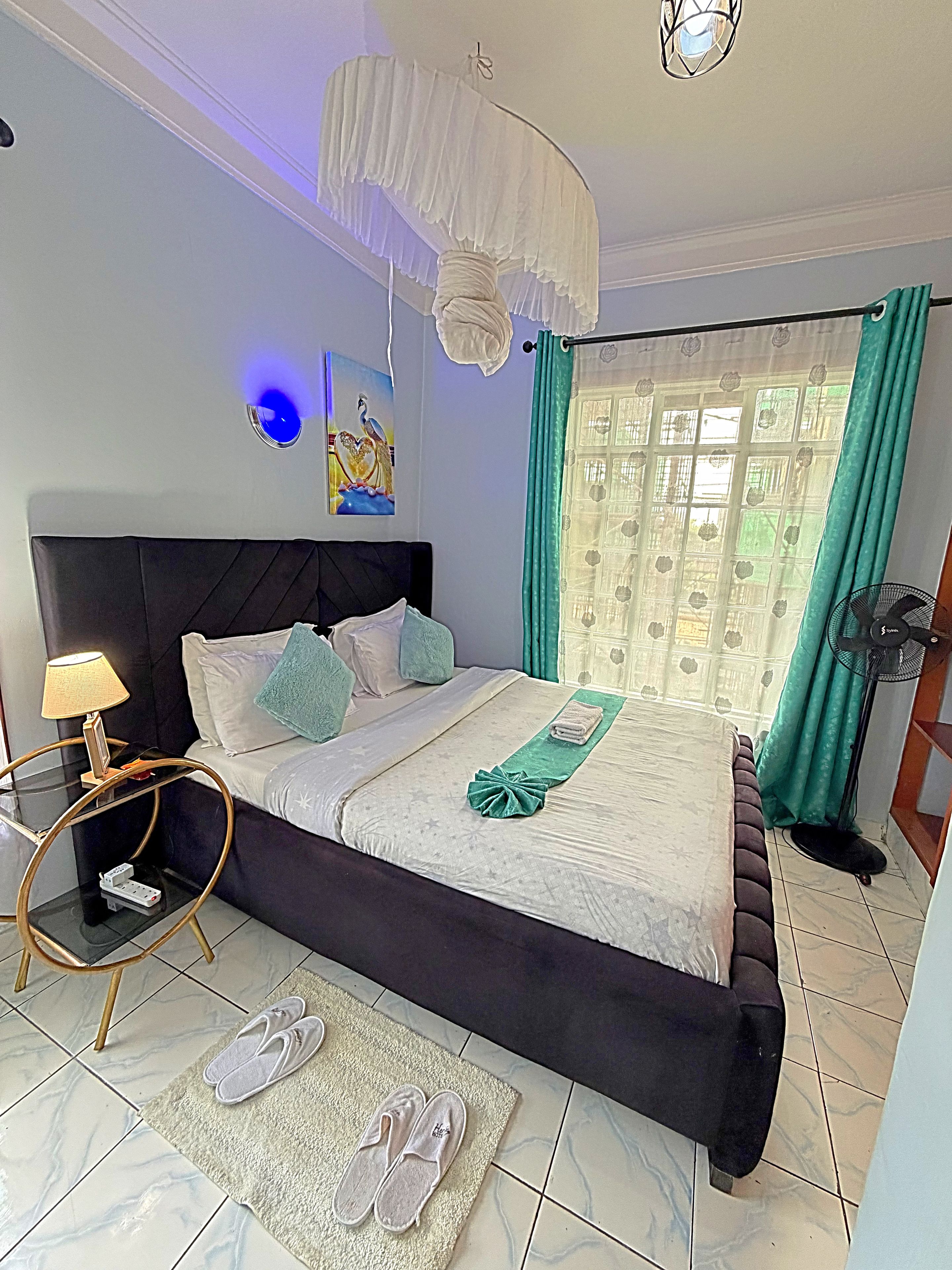 Ein Schlafzimmer mit einem Bett, einem Nachttisch mit Lampe, einem Ventilator und einem Paar Hausschuhen auf dem Boden.