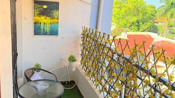 A balcony with a table and chairs, an air conditioning unit, a wall-mounted painting, and a view of greenery.