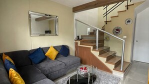 Una sala de estar con un sofá gris, una mesa de centro de vidrio y una escalera con escalones de madera y barandilla de vidrio.