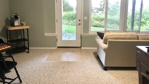 A living room with a beige sofa, a coffee machine on a wooden table, and a view of a garden through a glass door. - Water view high bank two bedroom private border Manchester State Park. (Port Orchard)