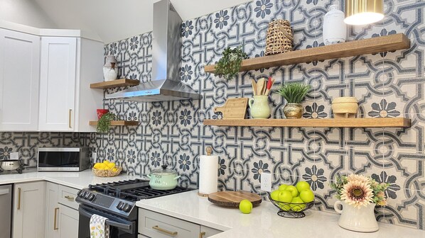Een moderne keuken met witte kastjes, een fornuis en een achterwand met een patroon. Er zijn houten planken met verschillende keukenbenodigdheden en een schaal groene appels op het aanrecht.