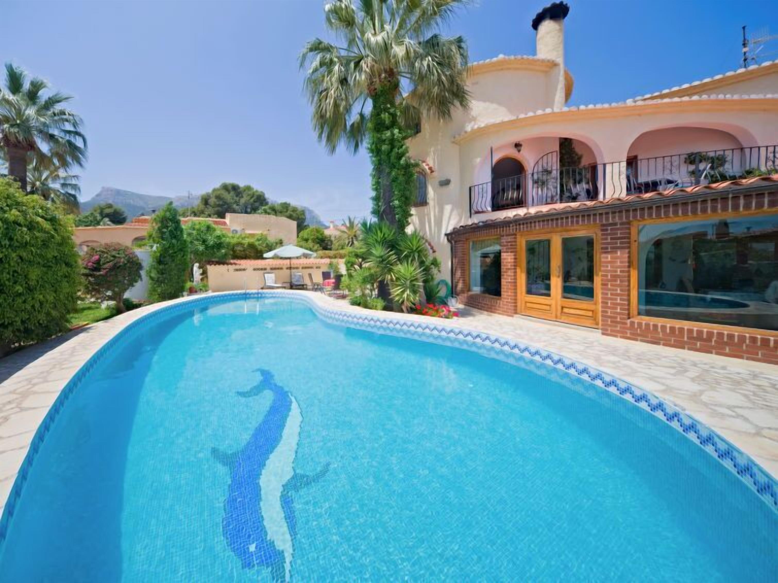 En swimmingpool med klart, blåt vand, omgivet af en fliseterrasse og en bygning med balkon.