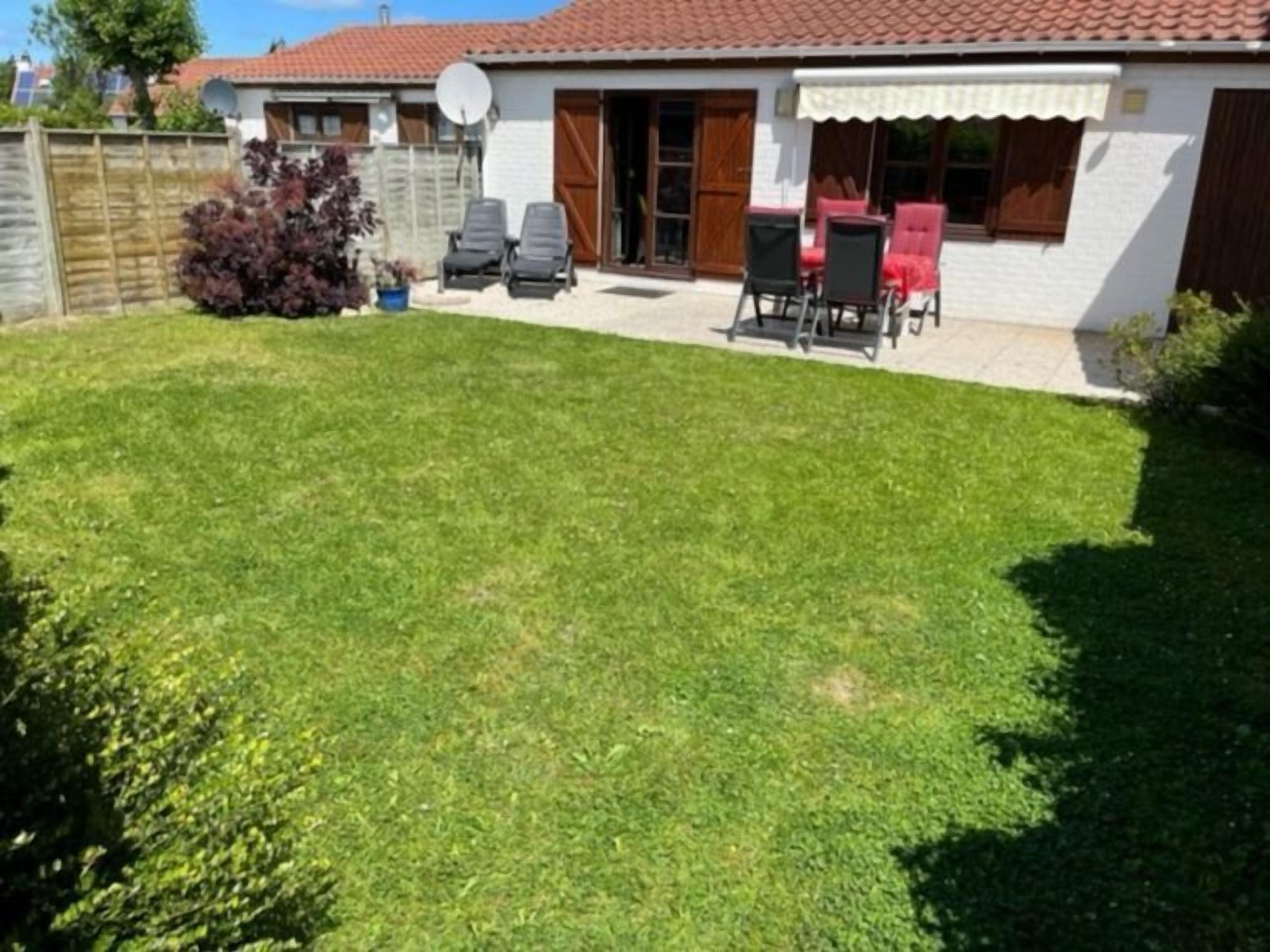 Una casa con techo de tejas rojas, fachada blanca y un pequeño patio trasero con césped y mobiliario exterior.