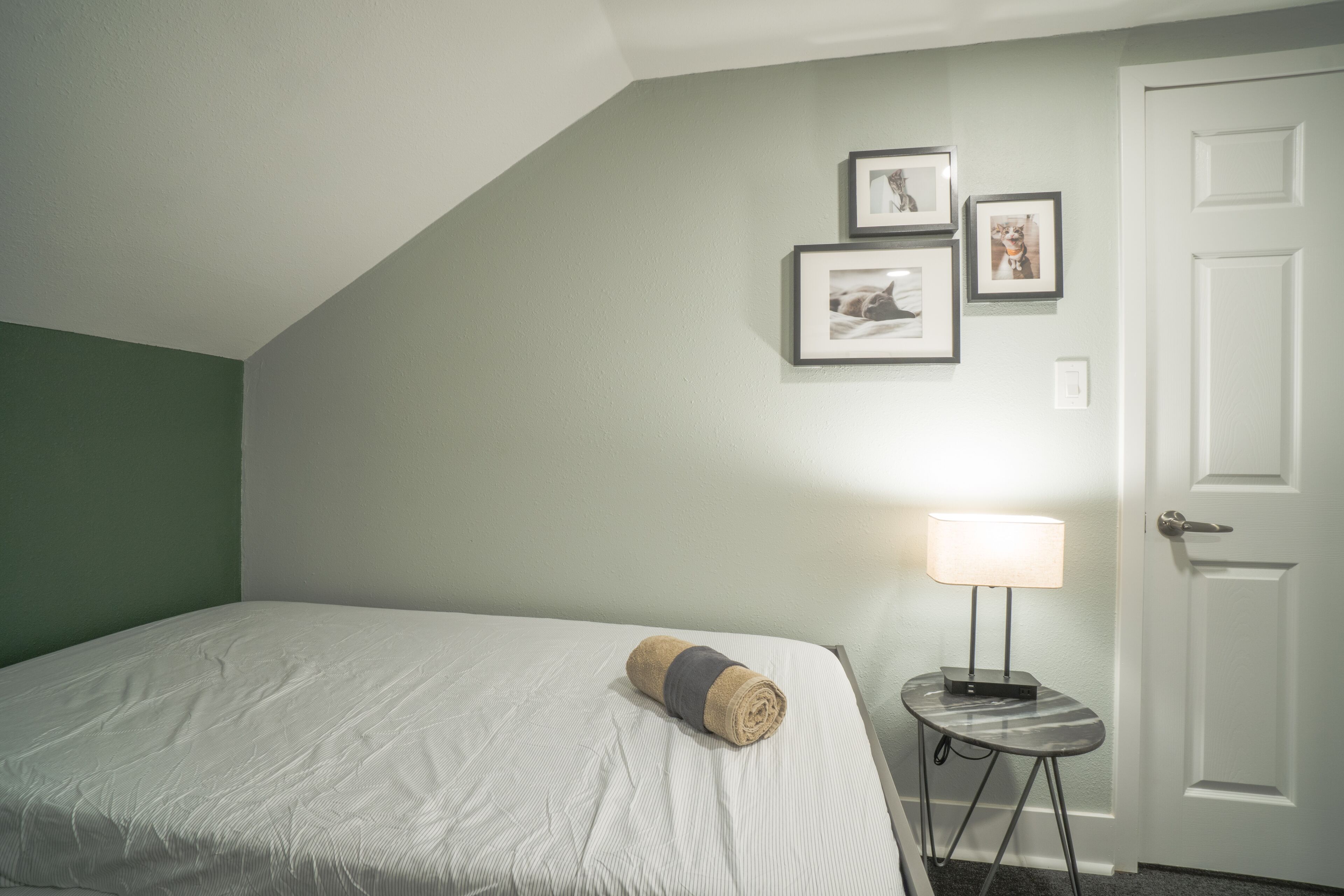 A neatly made bed with a rolled towel, a small table with a lamp, and framed pictures on the wall.