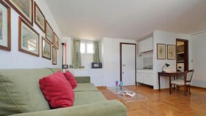 Ein Wohnzimmer mit einem grünen Sofa, einem Couchtisch und einem Essbereich mit Tisch und Stühlen. An den Wänden hängen Bilder in Rahmen und es gibt ein Fenster mit Vorhängen.