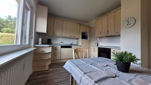 Una cocina con armarios de madera, una mesa de comedor con mantel, un reloj en la pared y una ventana con vistas a la vegetación.
