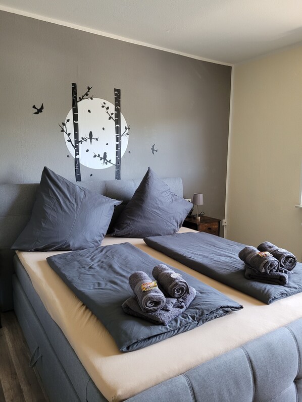 Un lit double avec une literie et des oreillers gris, deux serviettes enroulées posées sur le lit, une table de chevet avec une lampe, et un motif décoratif représentant des oiseaux et un arbre sur le mur.