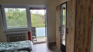 Un dormitorio con cama, radiador, armario de madera y puerta corredera de vidrio que da a un espacio exterior con una silla y una mesa.