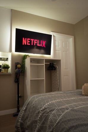 Ett sovrum med en säng, en garderob, en dammsugare och en väggmonterad tv som visar "NETFLIX".