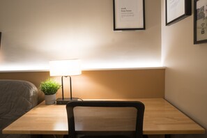 A hotel room with a wooden desk, a chair, a potted plant, a lamp, and framed pictures on the wall.