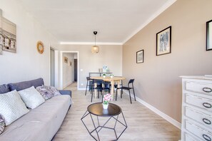 A living room with a sofa, a dining table with chairs, a small round table with a flower, and a dresser with drawers.