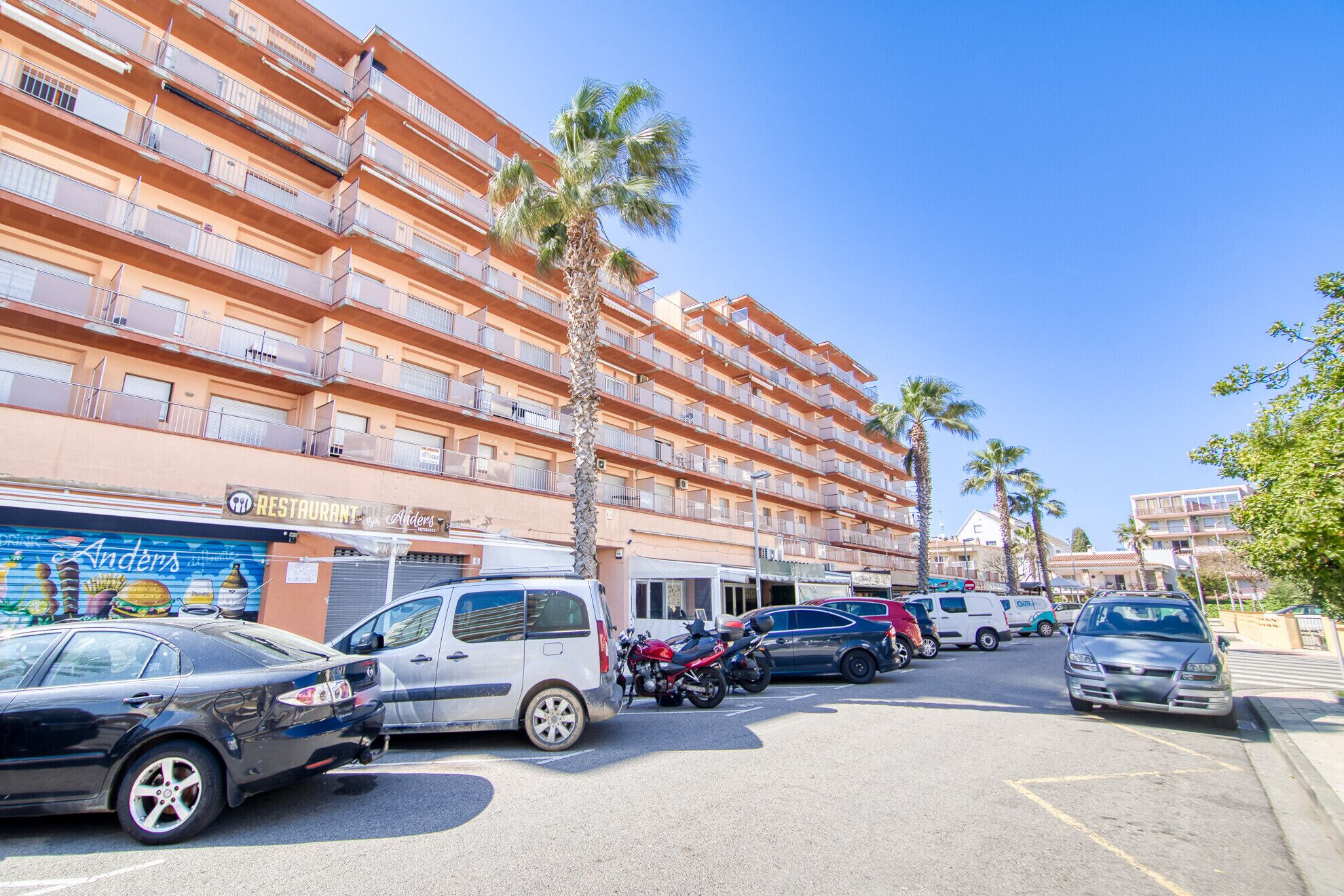 A multi-story building with balconies, parked cars, and palm trees.