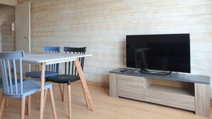 A modern living room with a wooden wall, a dining table with chairs, a flat-screen TV on a wooden stand, and a clock on the wall.
