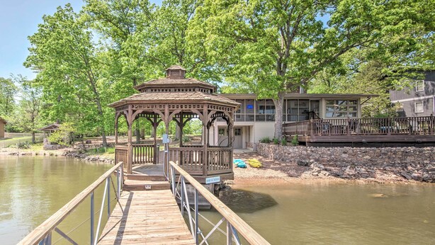 Eine Holzstege führt zu einem Pavillon an einem ruhigen Fluss, mit einem Haus und einer Anlegestelle im Hintergrund.