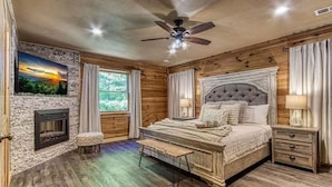 A bedroom with a stone fireplace, a large bed with a tufted headboard, a ceiling fan, and a view of greenery outside. - …STUNNING Luxury 3BD Lodge! …HOT TUB/Games/VIEWS!… (Pigeon Forge)