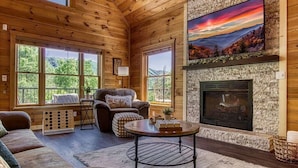 A cozy cabin interior with a fireplace, wooden walls, and large windows offering a view of the outdoors. - …STUNNING Luxury 3BD Lodge! …HOT TUB/Games/VIEWS!… (Pigeon Forge)