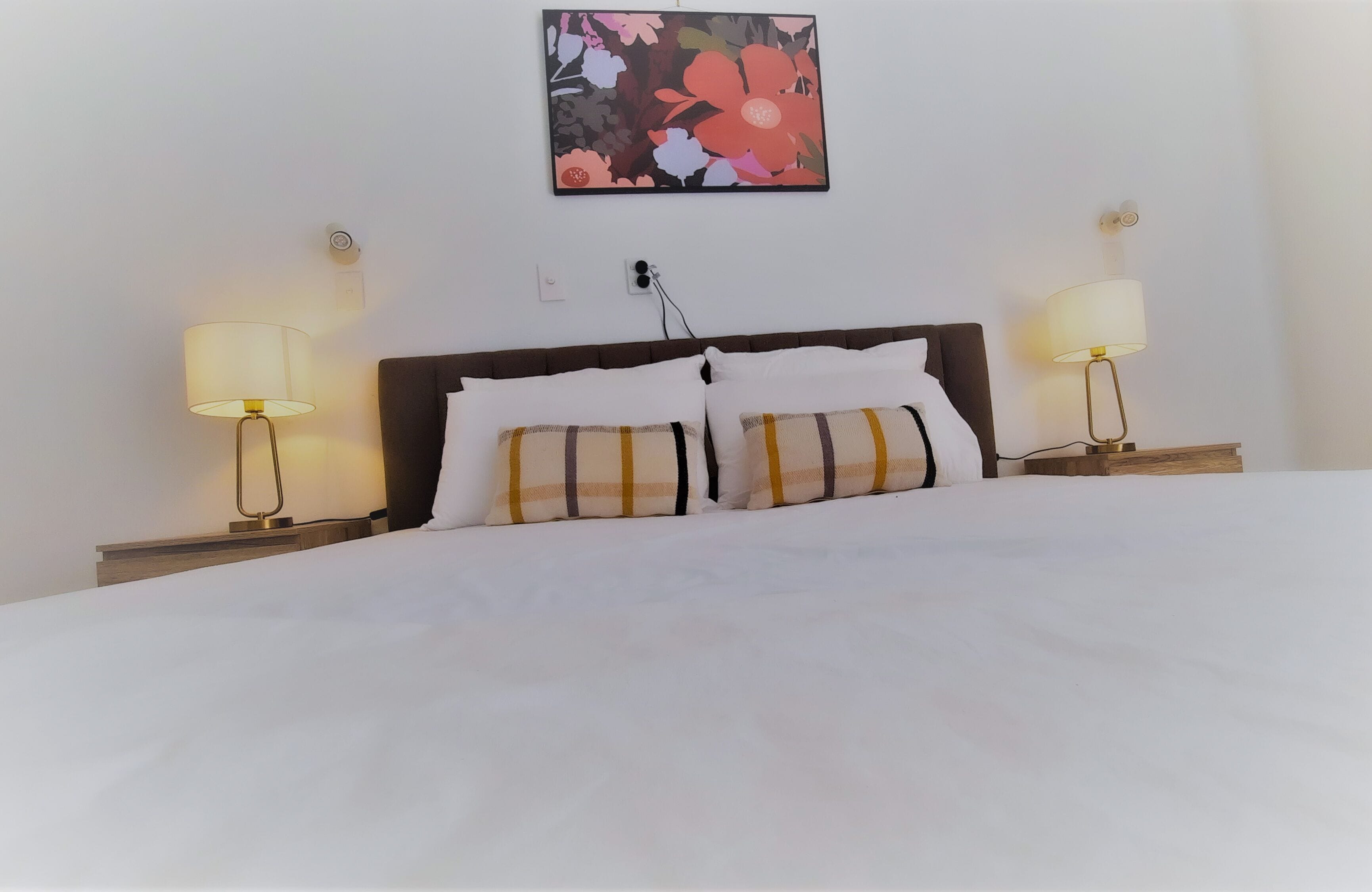 Cama bem arrumada com lençóis brancos e almofadas listradas, com duas mesas de cabeceira com luminárias, em um quarto com uma obra de arte floral na parede.