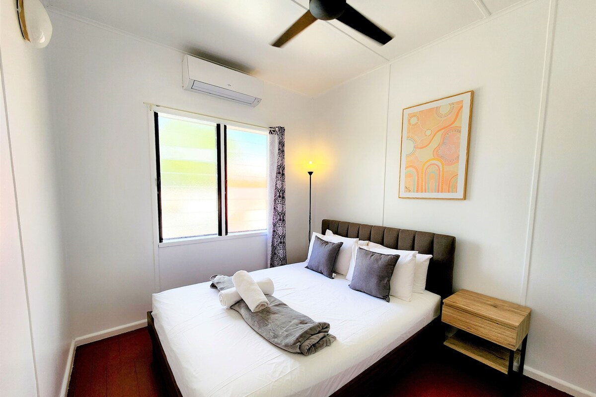 A neatly made bed with pillows and a rolled towel, a bedside table, a standing lamp, a ceiling fan, a window with curtains, and a wall-mounted artwork.
