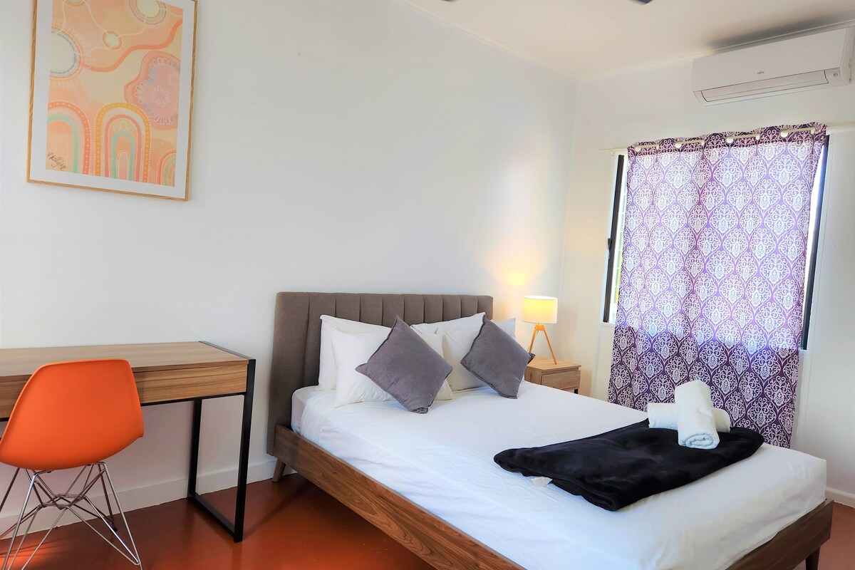 A bedroom with a bed, a desk, an orange chair, a ceiling fan, a window with patterned curtains, and a wall-mounted air conditioner.