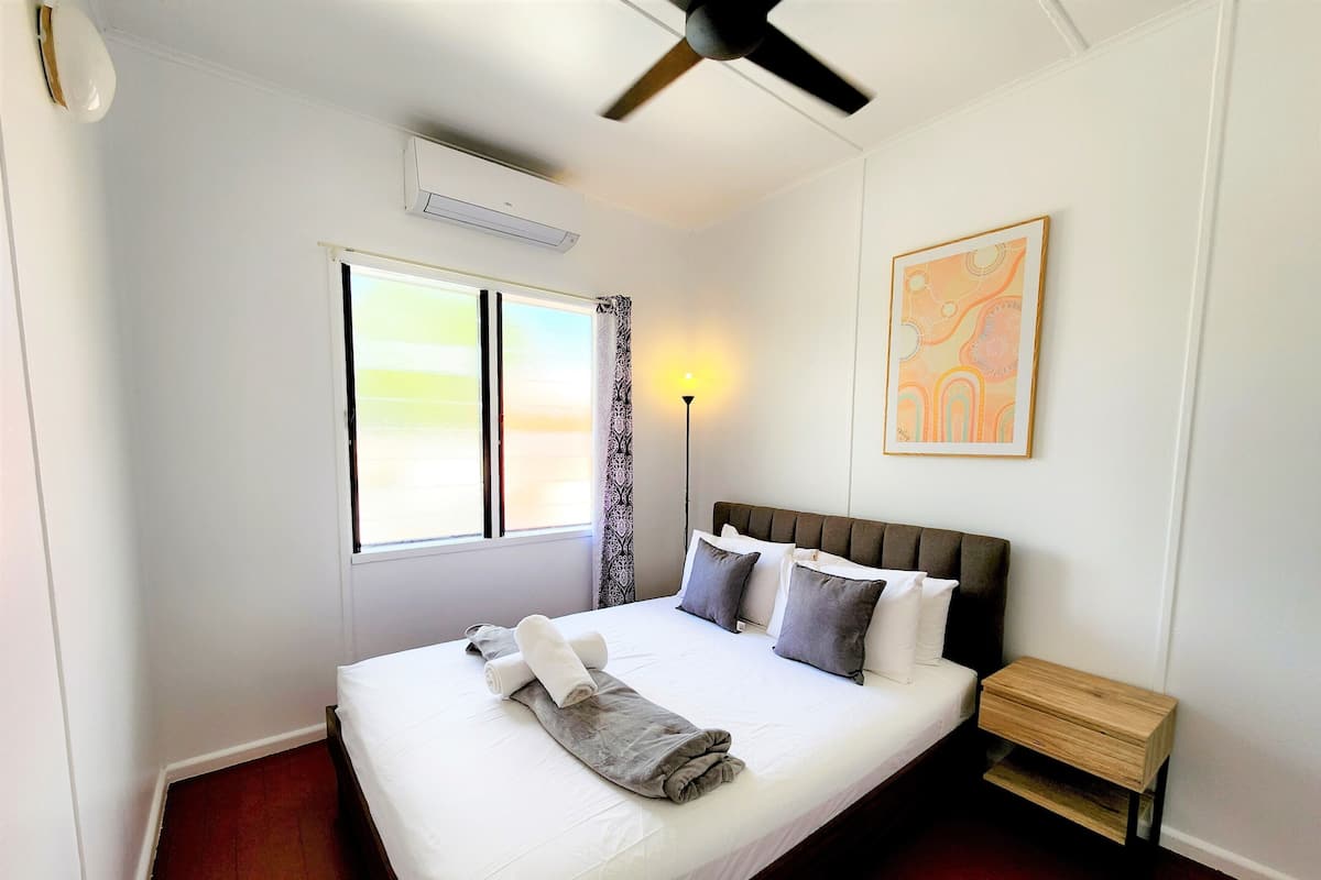 A neatly made bed with pillows and a rolled towel, a bedside table, a standing lamp, a ceiling fan, a window with curtains, and a wall-mounted artwork.