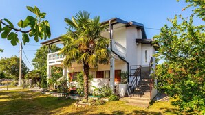 Una casa de dos pisos con balcón, escaleras y un jardín con diversas plantas.