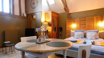 Een gezellige kamer met een houten eettafel, twee stoelen, een bed met kussens, een lamp en een klok aan de muur.