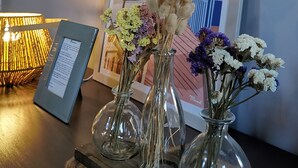 Un quadro con una locandina di 'CAP FERRET' appesa al muro, tre vasi di vetro con fiori secchi su un piano di legno e una lampada da tavolo.