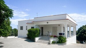 Una casa blanca de dos pisos con techo de tejas, un porche cubierto y un pequeño jardín con plantas en macetas.