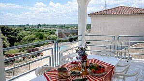 Una terraza con una mesa lista para una comida, rodeada de sillas blancas, con vistas a un paisaje con vegetación y un edificio a lo lejos.