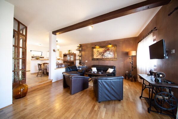 A spacious living area with wooden flooring, a dark leather sofa set, a dining table, and a television on a stand.