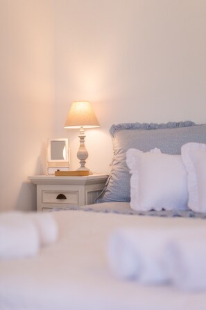 A neatly made bed with a white comforter and pillows, a bedside table with a lamp and a framed picture, and a wall with a light switch.