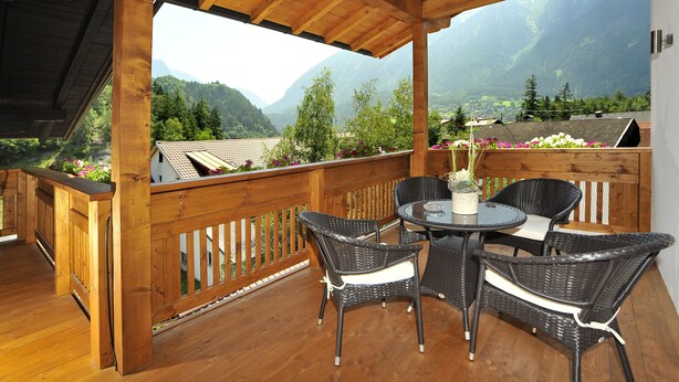 A wooden deck with wicker chairs and a table, overlooking a mountainous landscape with houses and greenery.