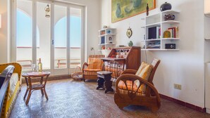 Ein gemütliches Wohnzimmer mit einem gemusterten Boden, einem Holztisch, Korbstühlen und einem Bücherregal mit Büchern und Dekoration.