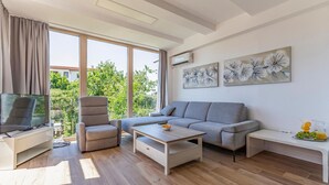 Uma sala de estar moderna com um sofá cinza, uma poltrona, uma mesa de centro e uma televisão. Há grandes janelas com cortinas, obras de arte na parede e vista para o verde lá fora.