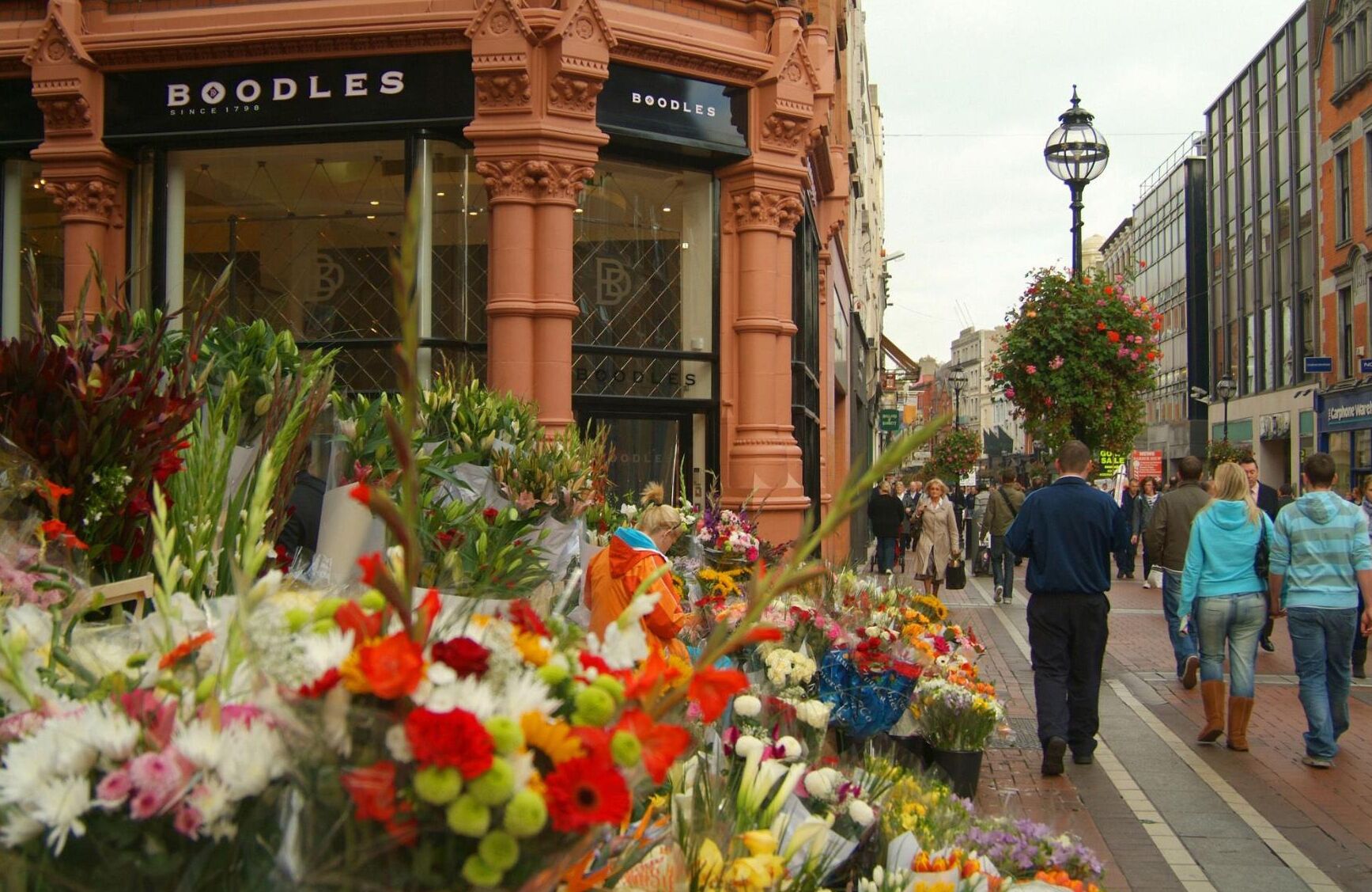Una calle con un mercado de flores y un edificio con letrero 'BOODLES'.