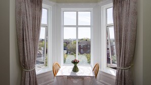 Ein Essbereich mit einem Tisch für zwei Personen, einer Blumenvase und Blick auf die Nachbarschaft durch ein Erkerfenster.