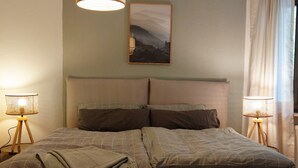 Una cama bien hecha con cabecera, dos lámparas de noche, un cuadro en la pared y una ventana con cortinas.