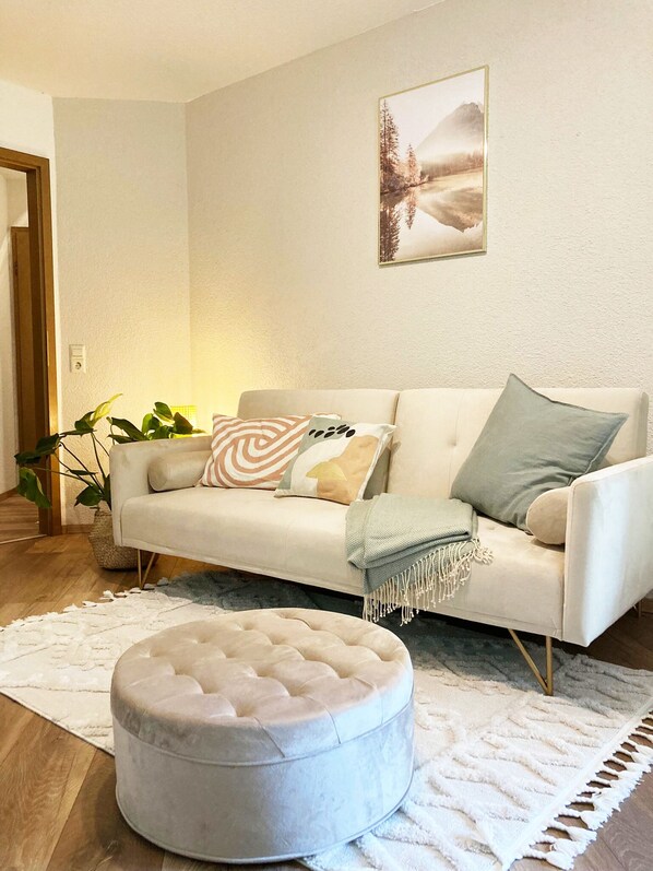 A living room with a white sofa, a round ottoman, a framed picture on the wall, and a plant.