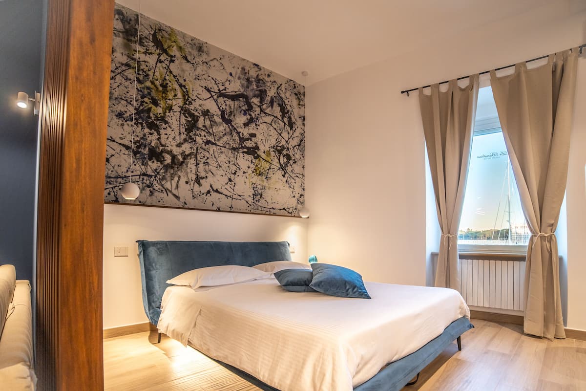 A modern bedroom with a large bed, a wooden floor, a ceiling artwork, and a window with curtains.