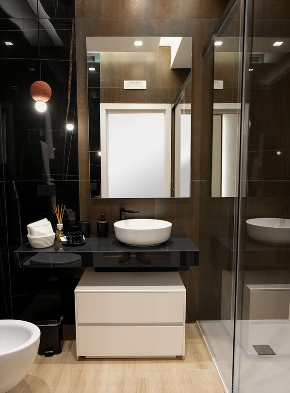 A modern bathroom with a white sink, dark tiles, a large mirror, and a glass shower enclosure. - LA BANCHINA LUXURY SUITES (Trani)