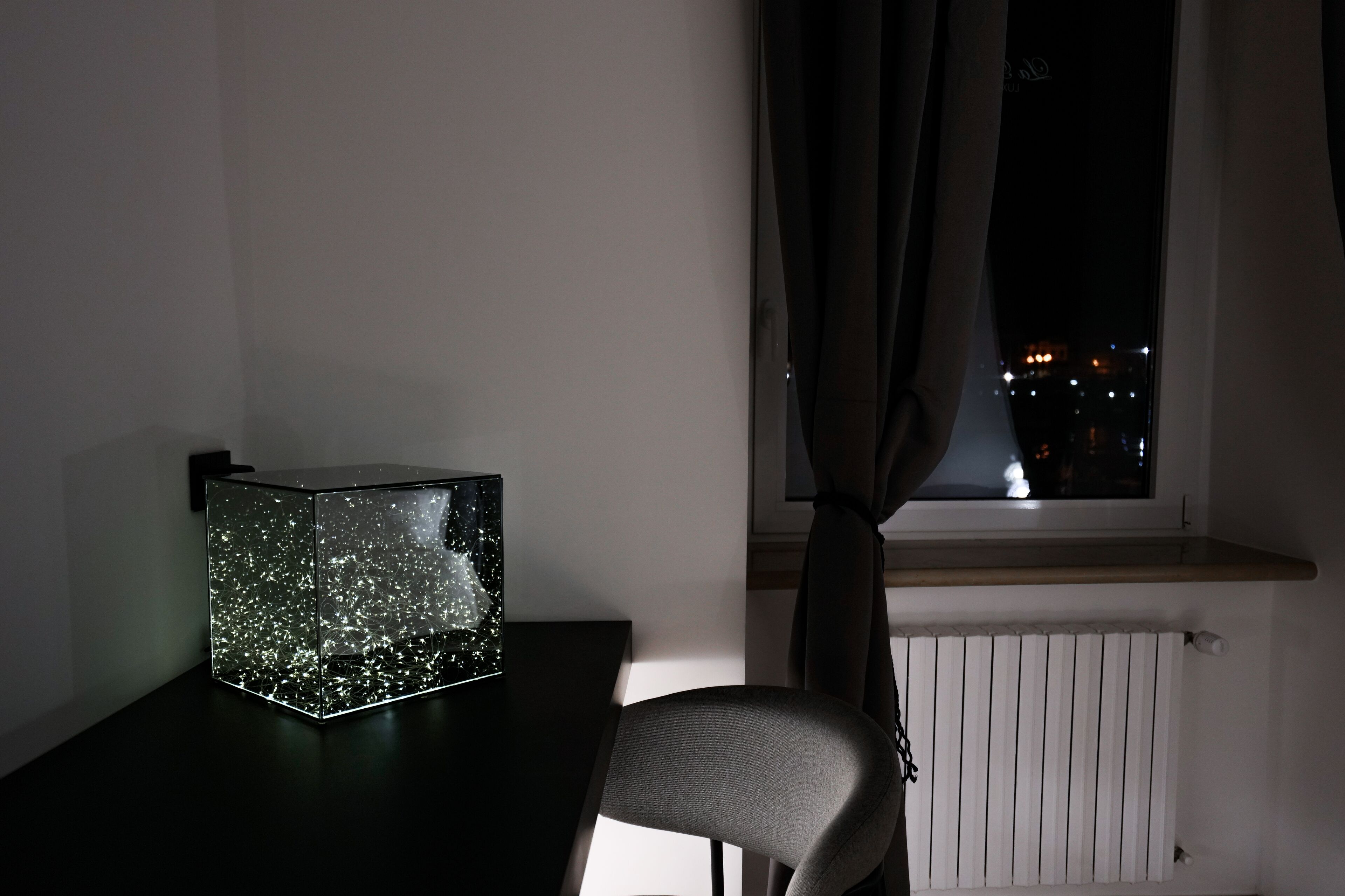 Una habitación con una mesa, una silla, una ventana con cortinas y una vista nocturna de la ciudad.