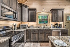 A kitchen with dark wooden cabinets, stainless steel appliances, a granite countertop, and a window with a view of trees.