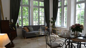 A living room with a sofa, a coffee table, and a chair. There are windows with curtains, a lamp, and a vase with flowers. - The Orangerie in the grounds of Chateau St Justin (Saint-Justin)