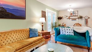 A living room with a large painting, a sofa, a teal sofa, a lamp, and a coffee table. - Lovely Home near the river and 5 mins to downtown AVL. (Asheville)