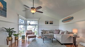 Ein geräumiges Wohnzimmer mit Deckenventilator, großem Fenster, grauem Sofa, Couchtisch und einem kleinen Beistelltisch.